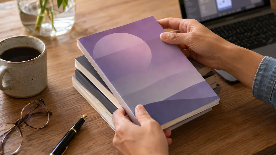 Hands placing a softcover paperback book onto a wooden desk next to a coffee cup, fountain pen, and reading glasses