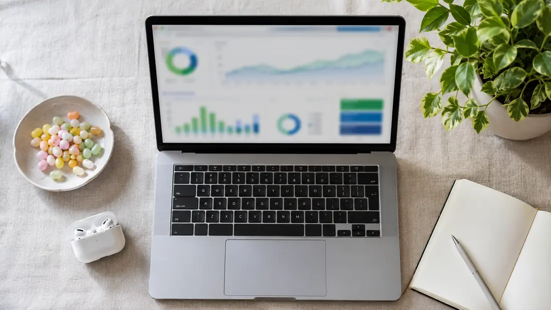 Modern overhead shot of a sleek silver laptop displaying analytics dashboards, beside earbuds, a notebook with pen, jellybeans in a dish, and a leafy plant on a linen surface