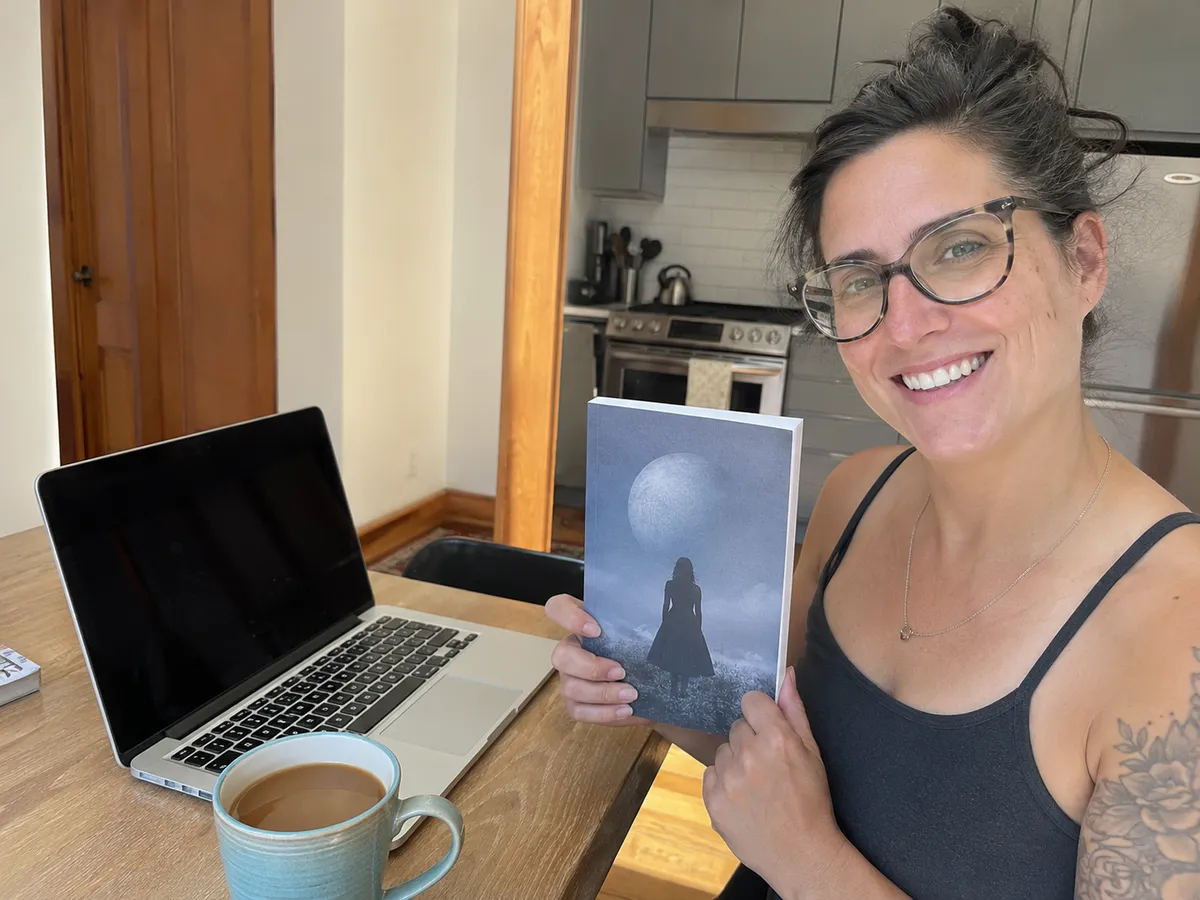An author sitting at a sunny kitchen table with an open laptop and a coffee, smiling and holding up a finished paperback she made with Inkfluence AI, the cover showing a moonlit figure on a dark blue background.