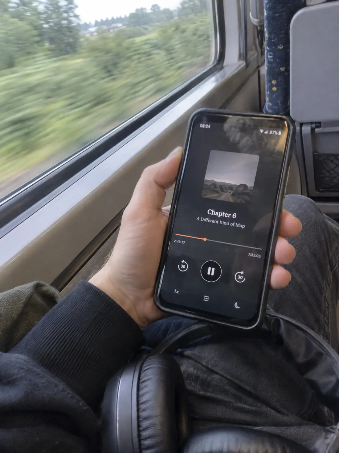 A commuter on a train holding a phone showing an audiobook player, with the elapsed and remaining time either side of the progress bar