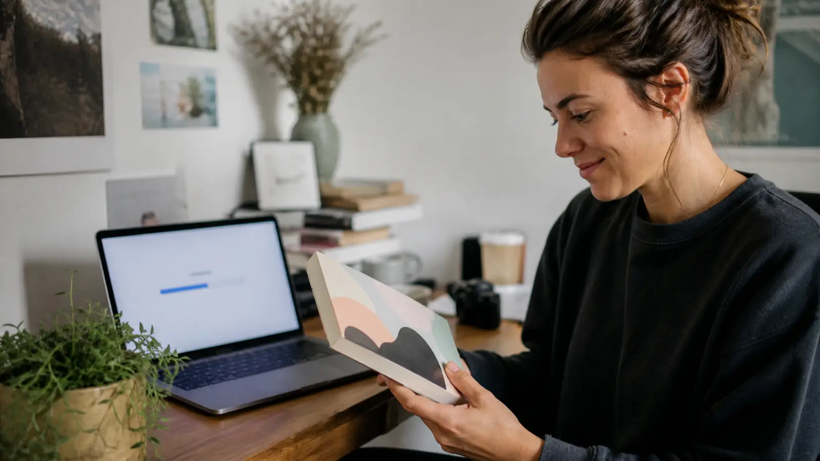 Author smiling at a printed paperback book with an abstract cover, laptop in soft focus showing an upload progress bar.