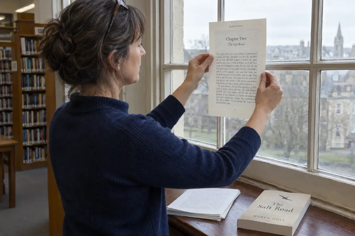 An author at a library window holds a printed page of The Salt Road by Maren Holt against the glass to check that the text blocks on the front and back of the sheet line up, with the finished paperback on the sill
