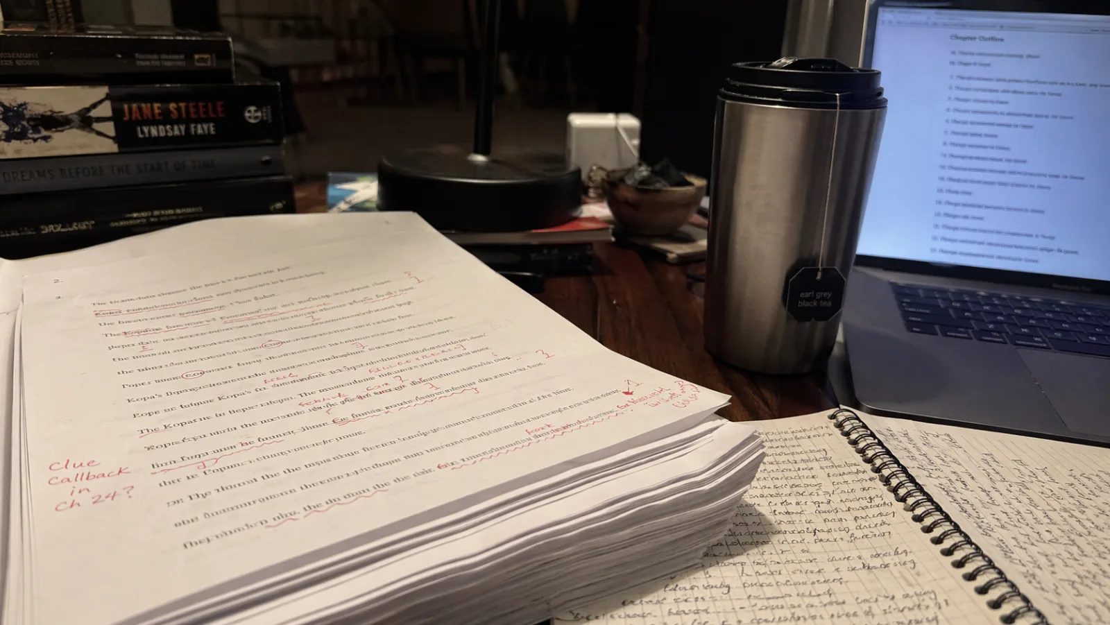 A novelist's printed manuscript covered in red-pen edits, circled passages, and handwritten margin notes reading 'clue callback in Ch 24?', with a thermos of tea and a scruffy notebook beside the stack and a thin laptop blurred in the background showing a chapter outline, photographed under warm overhead lamplight at night during a revision session.