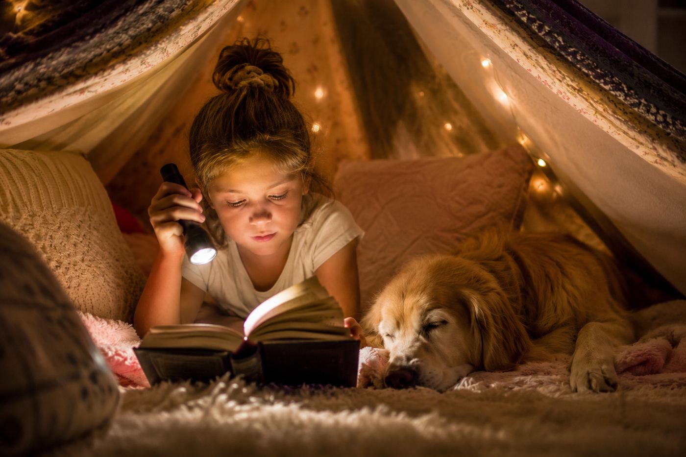 A proud seven-year-old reading a printed chapter book alone under a blanket fort with a flashlight