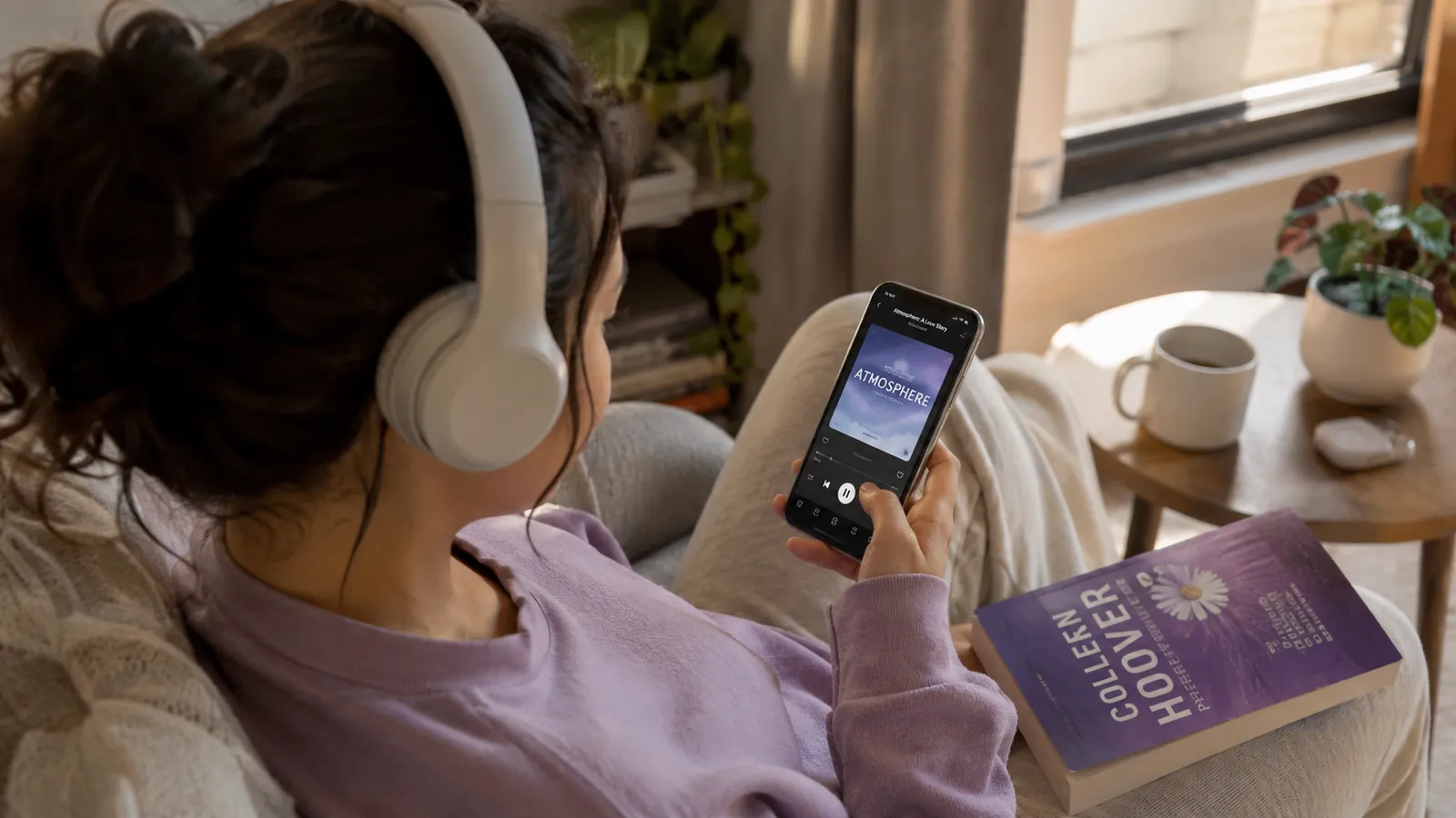 A person relaxing on a sofa with over-ear headphones, listening to an audiobook on their phone with a paperback beside them