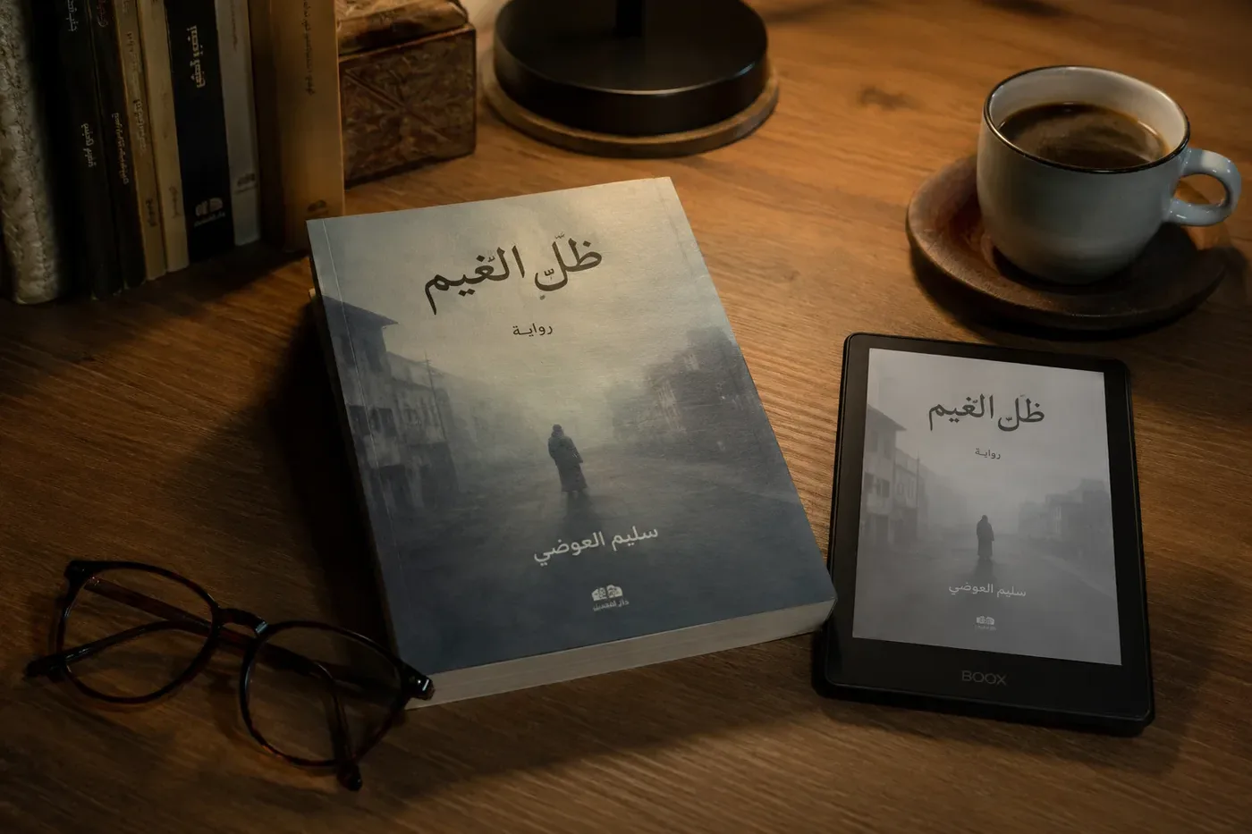 طاولة قراءة عليها رواية عربية مطبوعة بغلاف طباعي هادئ، وبجانبها قارئ إلكتروني يعرض غلاف الرواية نفسها، ونظارة قراءة وفنجان قهوة، في ضوء مصباح دافئ.