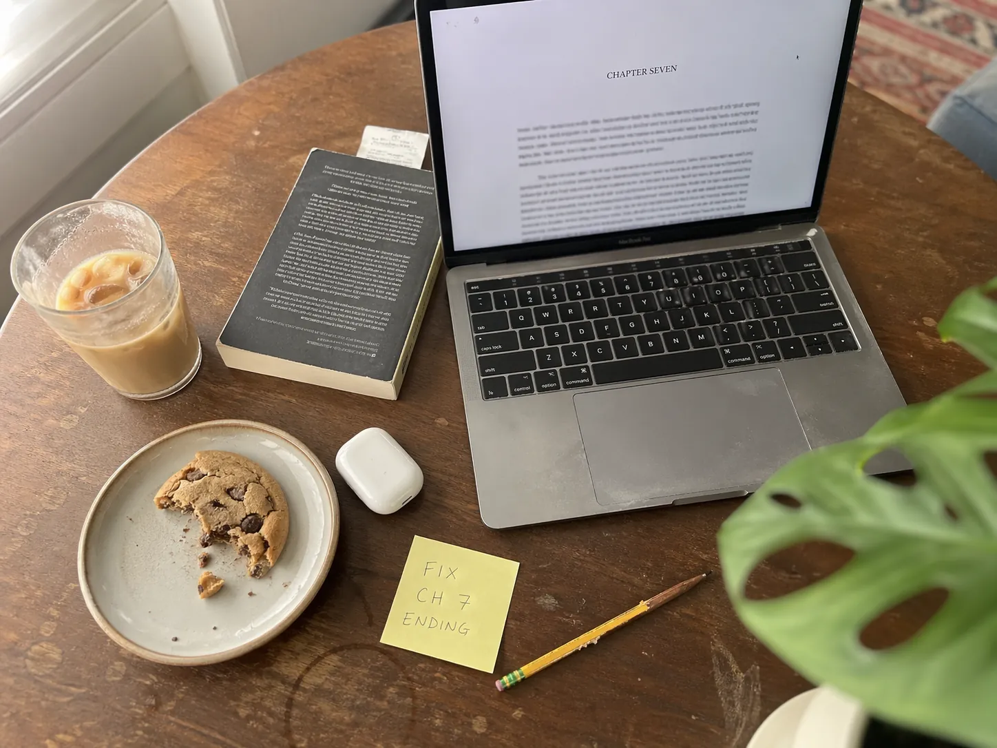 Writer's desk mid-revision with a MacBook open to Chapter Seven of a draft novel, an iced oat-milk latte, a half-eaten chocolate chip cookie, AirPods, a yellow Post-it note that reads 'FIX CH 7 ENDING', a chewed pencil, paperback bookmarked with a receipt, and a monstera leaf in soft focus