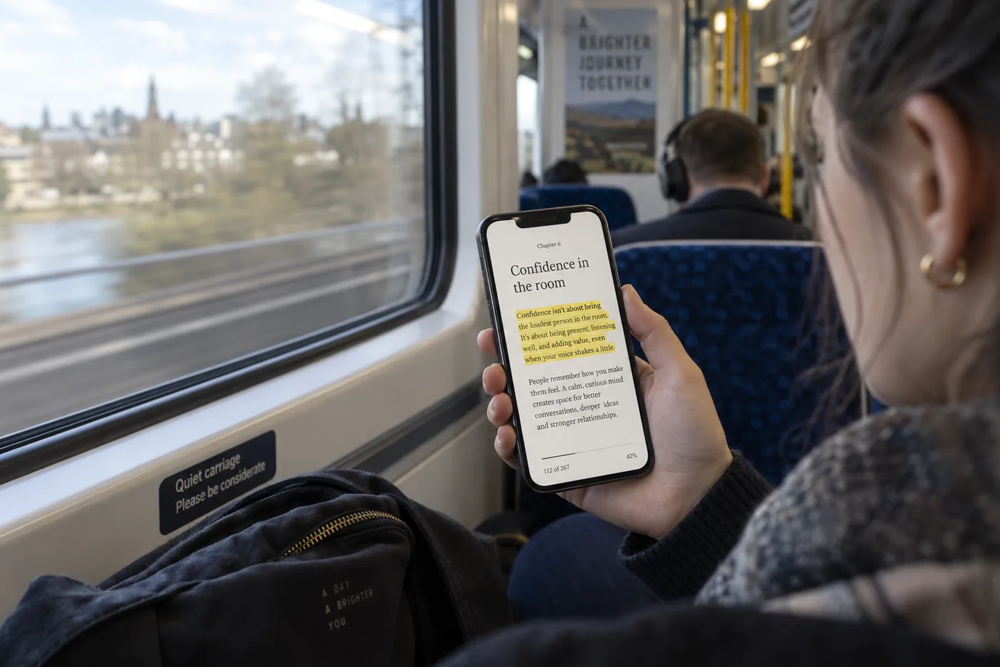 A reader on a train with a phone showing a self-help ebook open at a chapter on confidence, a highlighted passage visible.