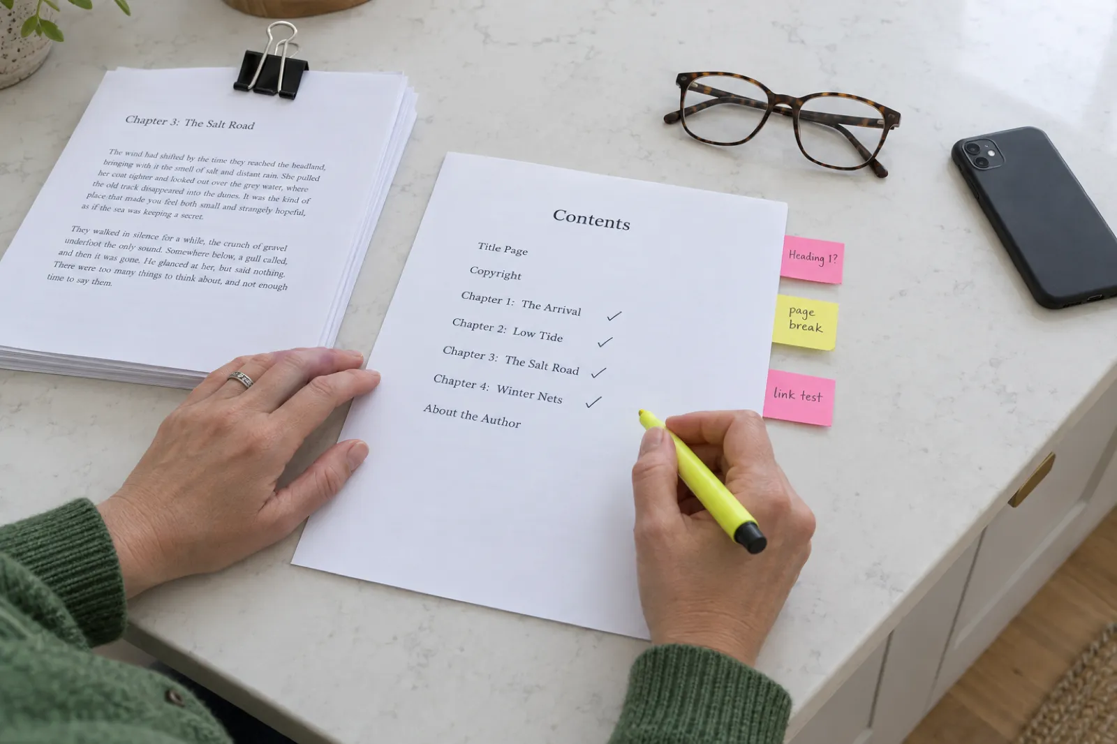 A woman in a green cardigan highlights a printed Contents page listing Title Page, Copyright, Chapter 1: The Arrival, Chapter 2: Low Tide, Chapter 3: The Salt Road, Chapter 4: Winter Nets and About the Author, each chapter ticked, with sticky notes reading Heading 1?, page break and link test along the edge, beside a clipped manuscript and reading glasses.