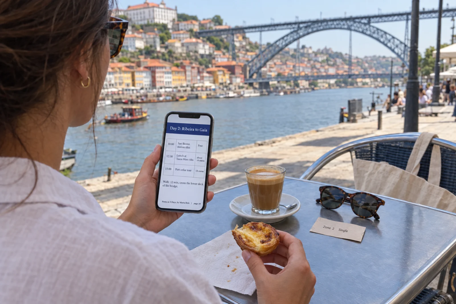 Over the shoulder of a woman at a cafe table on the Porto riverfront, the iron Dom Luis I bridge behind her, reading a travel guide PDF on her phone at a page headed Day 2: Ribeira to Gaia with a table of stops: 10:00 Sao Bento station tiles, free; 12:30 lunch at Tasca Mare Alta, about 14 euros; 15:00 port cellar tour, 18 euros, and the line Walk: 12 min, cross the lower deck of the bridge, with the footer Porto in 3 Days, by Marta Reis, page 14; a custard tart in her other hand, a cortado, sunglasses and a paper tram ticket on the table
