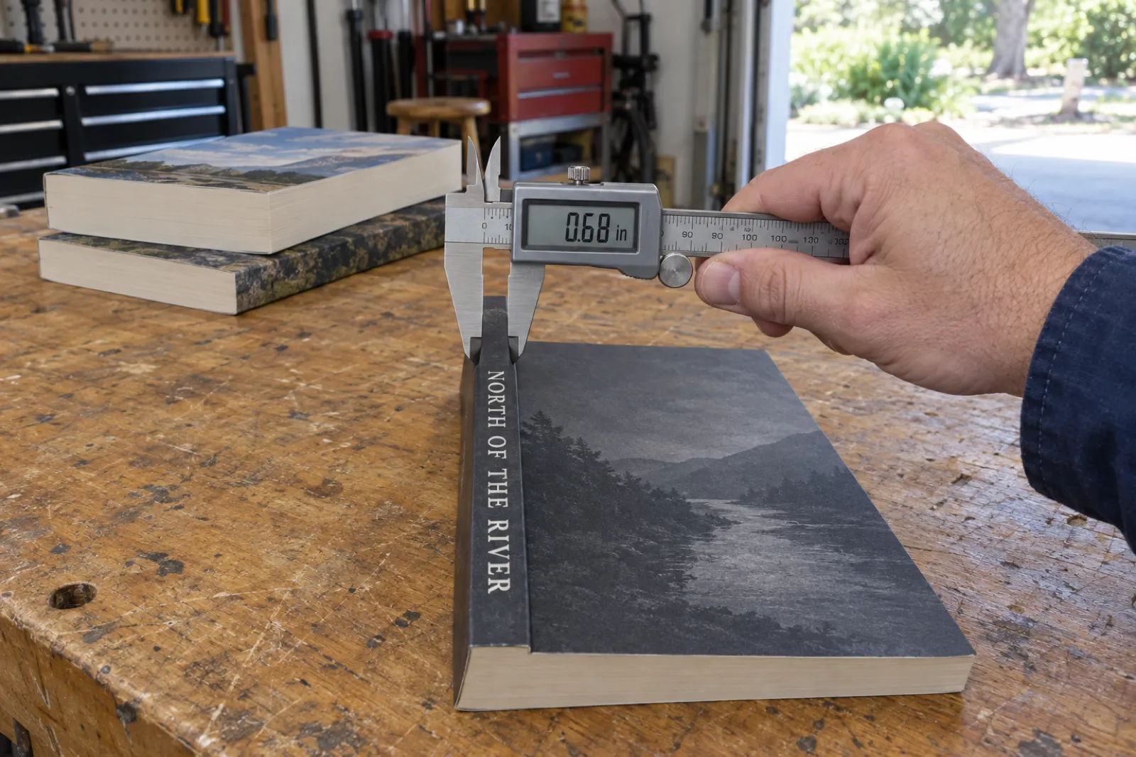 A hand holds a digital caliper reading 0.68 in against the spine of a charcoal 6 by 9 inch paperback titled North of the River on a garage workbench, with two thicker paperbacks stacked behind.