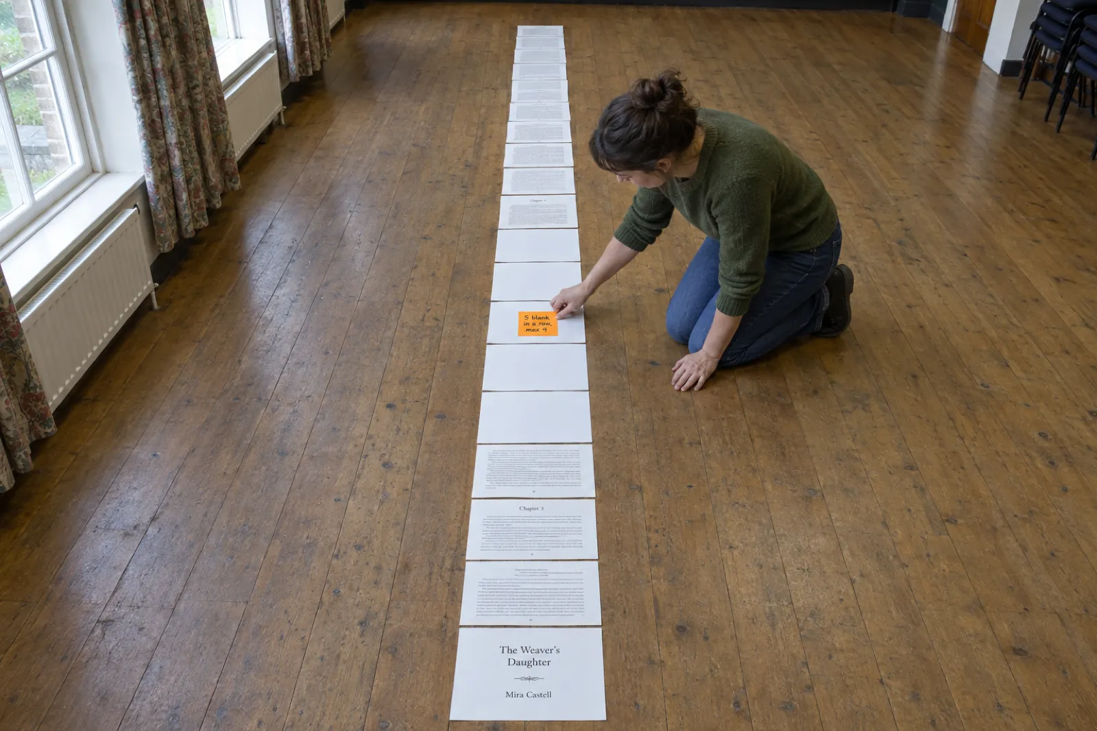 A printed book interior of The Weaver's Daughter by Mira Castell laid out as one long line of pages on a village hall floor, with a run of five blank pages between Chapter 3 and Chapter 4 and a woman pressing on an orange sticky note reading 5 blank in a row, max 4